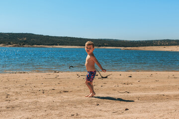 A happy little boy stands on the beach of the reservoir with a branch in his hands and looks at the camera.