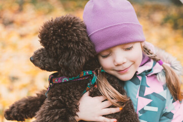 Portrait of a little cute girl, closing her eyes, embracing a poodle dog in an autumn park of yellow fallen leaves.