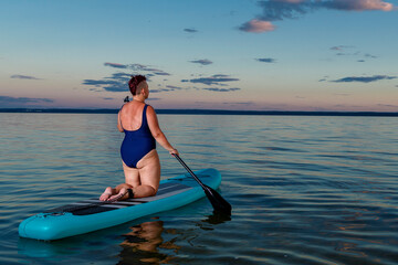 A woman with a short Mohawk haircut in a swimsuit on a SUP board with a paddle floats on the water.