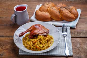 Spelled with vegetables and a duck leg in a light plate next to it on a napkin on the table next to the challah and a jug of wine.