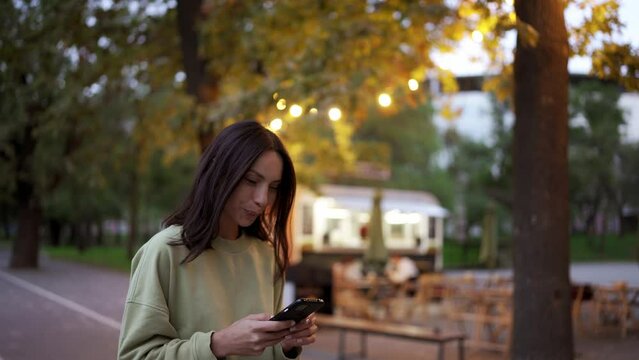 Brunette girl in a green sweater is chatting on the phone against the backdrop of an evening park. Cafe in the park, spending time in the city. An evening walk