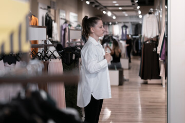 Fototapeta na wymiar A young pretty woman in white clothes buys clothes in a boutique store, tries on a white shirt at the mirror , lifestyle real life photo clothing selection new collection