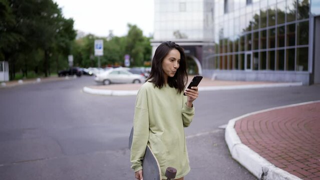 A brunette girl in a green sweater walks in the park. The girl has a skateboard in one hand and a phone in the other. The girl is texting on the phone. Evening walk