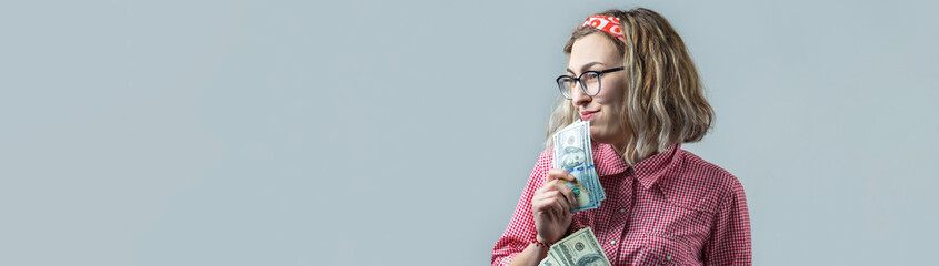 Closeup of young beautiful woman in a red plaid shirt in glasses with american dollars money in hand over gray background.