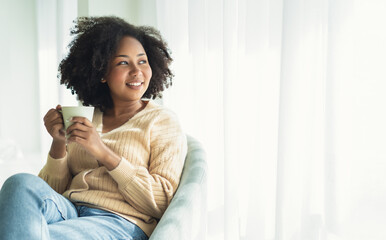 Beautiful young African American woman relaxing on comfortable chair and enjoying a cup of coffee...