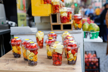 Fruit slices in transparent plastic cups with forks on a street market counter. Refreshing bright fruits on a hot summer sunny day.