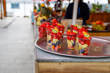 Fruit slices in transparent plastic cups with forks on a street market counter. Refreshing bright fruits on a hot summer sunny day.
