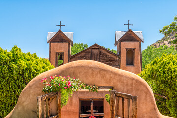 Naklejka premium Famous historic adobe El Santuario de Chimayo sanctuary church in the United States with entrance gate closeup of flowers in summer