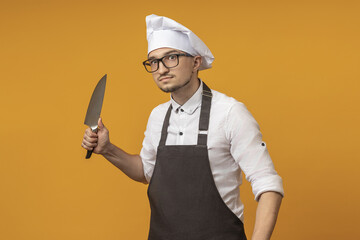 a young chef man in a black apron, beret and glasses stands with knives in his hands against a colored background