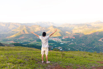 Man on top of a mountain with arms raised looking at the view with valley and high mountains in the background. Man experiencing freedom, success and happiness on top of a mountain.