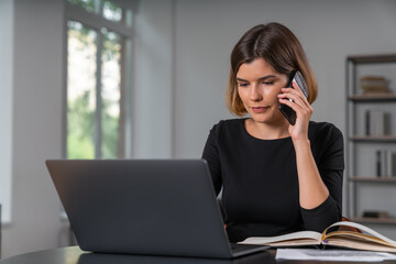 Pleased attractive businesswoman typing on laptop talking on smartphone