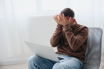 Unexpected error on computer device concept. Crying suffering upset young man closes his face with both hands lost important data due to laptop breakdown sitting on chair at home. Copy space