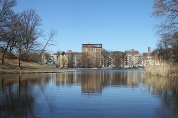 Landscape of buildings reflecting on Harlem Meer lake with trees under blue sky