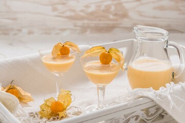 Two glasses and a carafe with eggnog and physalis fruits on a white wooden tray on a white marble...