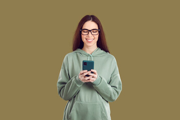 Satisfied pretty young Caucasian woman holding mobile phone in front of using dating apps or social networks to communicate with boyfriend wearing casual sweatshirt stands in green studio