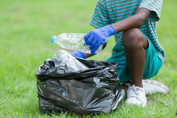 Volunteer kid. African American boy in gloves picking up plastic bottles into a black garbage bag outdoor in the park. Child boy picking garbages outside in the garden. Volunteer and charity concept