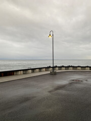 Lampadaire solitaire au bout d'un quai lors d'une journée nuageuse et grise. Berge d'un port et vagues du fleuve au loin à l'horizon.