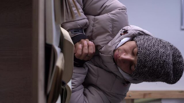 Student dressed in a warm winter hat and jacket sits stady with book in the hostel. Concept of no heating in winter at home.