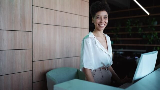 A smiling woman receptionist stands at the front desk and looking at the camera of the hotel