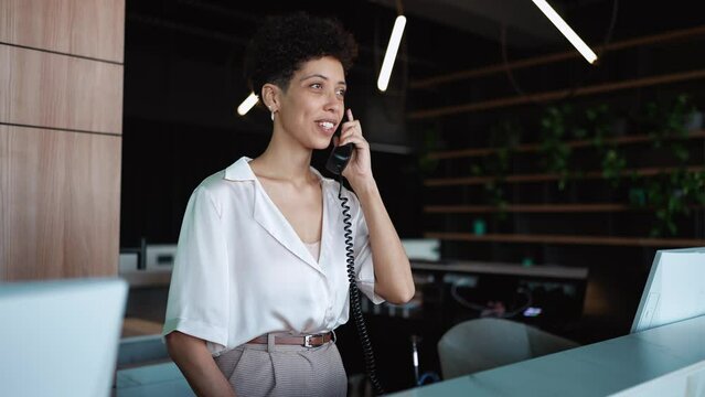 A cheerful woman receptionist stands at the front desk and talking on telephone of the hotel