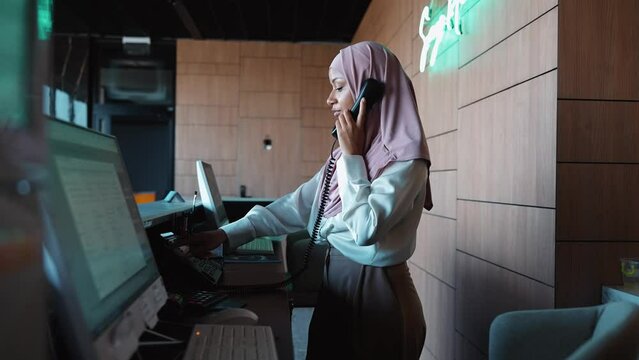 Positive Muslim woman receptionist stands at the front desk and talking on telephone of the hotel