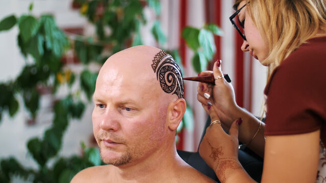 A girl, tattoo master, mehendi artist makes drawing of henna tattoo on scalp of bald Caucasian man, shoulder, neck.The solution of henna dries on the skin. High quality photo