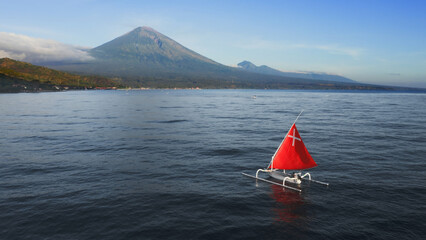 Fabulous scarlet red sail white Jukung boat floating on quiet ocean water near sea shore with huge volcano in summer. landscape with bright red sailboat swaying on wavy surface of sea Soft focus Pixel