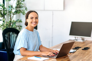 Photo of a successful confident gorgeous mixed race woman, business mentor, financial advisor, programmer, seo, sitting at a work desk in a modern office, looking at camera, smiling friendly