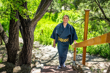 Young happy smiling man in kimono costume walking, leaning on railing fence in outdoor garden in...