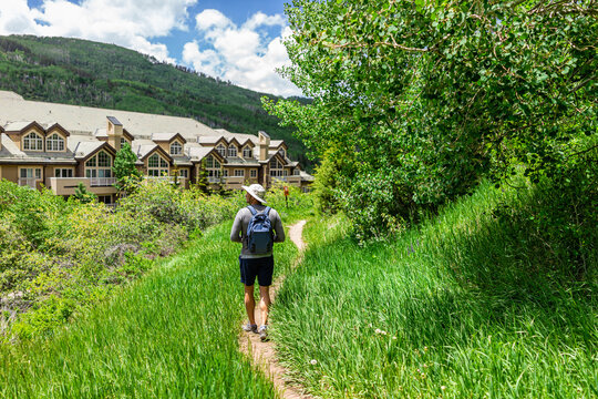 One young man back with backpack and hat walking on hiking trail of village loop in Beaver Creek ski resort near condo buildings in Colorado in summer