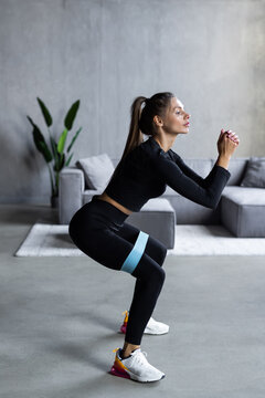 Home sports concept. Young woman doing squats with elastic band in living room