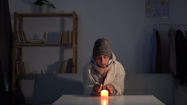 Freezing young man in winter clothes warms her hands on candle, energy blackouts cause electricity outages. energy crisis