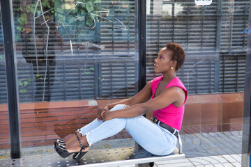 African American woman in a pink t-shirt, jeans and black heels, sitting at a bus stop with...