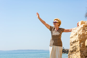 Elderly attractive smiling woman standing on a rock on the seashore waving her hand happily and enjoying life