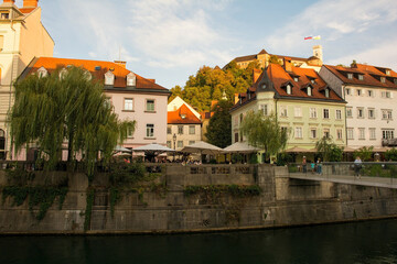 Fototapeta na wymiar Restaurants and bars on the waterfront of the Ljubljanici River in central Ljubljana, Slovenia. The Fishmarket Footbridge is foreground right, and the castle tower background right 