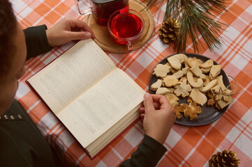 Naklejka na ściany i meble Overhead view of woman reading book, with blurred pages, while a tea time with fresh baked homemade gingerbread cookies and hibiscus hot drink. Christmas mood. Atmosphere of winter weekend or holidays