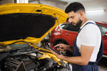 Serious focused man car technician mechanic repairing car problem of engine, during system checking detail, using tablet computer for maintenance and fixing in car garage.