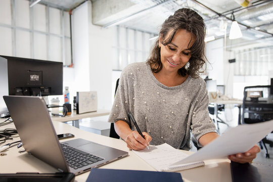 Smiling businesswoman with paperwork working at laptop in office