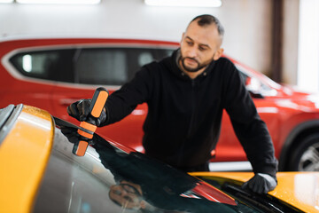 Male worker in black uniform and protective gloves, on a professional car wash service, applying anti rain coating on a windshield of luxury yellow car.