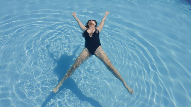 Woman wearing black swimsuit laying on surface of swimming pool as star fish. Girl with white skin floating on water with texture of ripples and sundog outdoors in summer. Spa hotel moments Soft focus