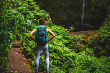 Naklejka na ściany i meble Athletic woman marvels at amazing jungle water fall with beautiful flowers and farns. Levada of Caldeirão Verde, Madeira Island, Portugal, Europe.