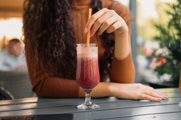 A girl with beautiful curly hair is stirring the freshly squeezed red juice in the cafe.
