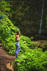 Naklejka na ściany i meble Sporty woman marvels at nature at an impressive waterfall in the jungle with beautiful flowers and ferns. Levada of Caldeirão Verde, Madeira Island, Portugal, Europe.