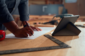 Close-up of young man holding pencil while leaning on paper opposite digital tablet in woodworking...