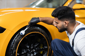 Washing car by hand, car detailing. Close up image of professional male worker, cleaning the car wheels and tires with brushes. Car rims tires wash, manual wheel cleaning concept.