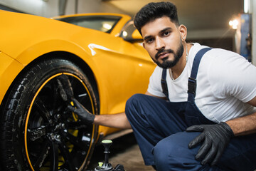 Fototapeta na wymiar Professional male worker in overalls and black protective gloves, cleaning alloy wheels rims of luxury car with a special brush for cast wheels in a detailing workshop.