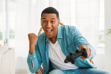 Satisfied excited young african american male with joystick playing online game, rejoices to win,...