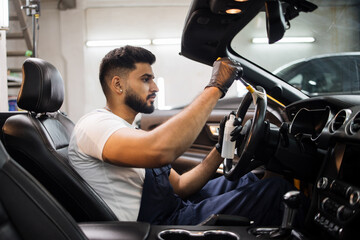Fototapeta na wymiar Car detailing and interior care concept. Young man, car wash worker, wearing protective rubber gloves, cleaning the steering wheel of the modern vehicle, using special brush.
