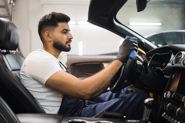 Obraz premium Handsome bearded young man in uniform and protective gloves, cleaning car interior and steering wheel using microfiber cloth, smiling at camera. Car detailing and valeting concept.