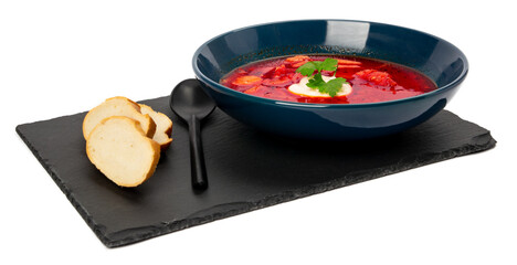 Ukrainian red soup borscht decorated with cream and herbs in center of dish on black board of stone with black spoon and pieces of white bread on white background isolate